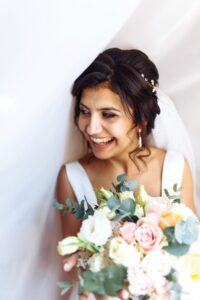 Young bride in white dress with a bouquet of flowers posing under curtain. Morning of the bride.