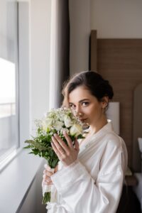 young bride with engagement ring on finger standing in white silk robe and smelling bridal bouquet